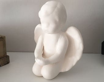 Może przedstawiać: Biała ceramiczna figurka anioła w pozycji klęczącej ze złożonymi dłońmi i dużymi skrzydłami. Anioł ma gładkie, matowe wykończenie i jest umieszczony na białej powierzchni.