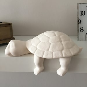 Op de afbeelding: Een mat wit keramieken schildpadbeeldje ontworpen om op de rand van een plank te zitten. De schildpad heeft een getextureerd, gesegmenteerd schild, een gladde kop en twee voorpoten die over de rand hangen in een rustende houding.