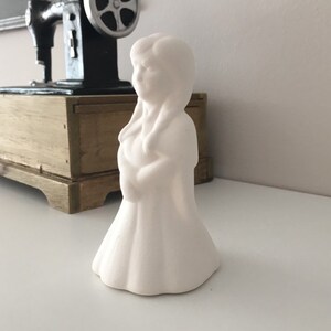 Puede incluir: Figurilla de cerámica blanca de una niña con el pelo largo, vestida con un vestido y de pie sobre una superficie blanca.