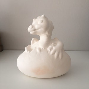 May include: White ceramic figurine of a baby dragon hatching from an egg. The dragon is partially out of the egg, with its head and front legs visible. The egg is cracked and has a rough texture.