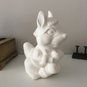 May include: A white ceramic squirrel ornament with a detailed design. The squirrel is seated, holding an object in its paws. The ornament has a smooth, unpainted surface, ready for painting. The squirrel has a detailed face and ears.