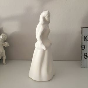May include: White ceramic figurine of a woman in a long dress. The figurine is standing on a white surface.