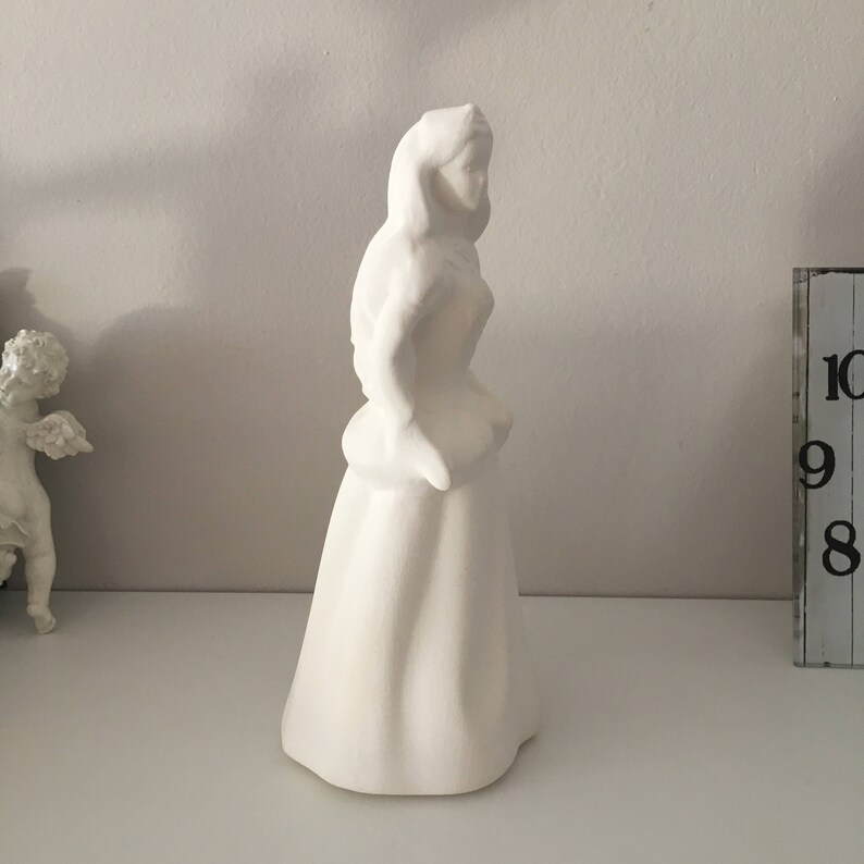 May include: White ceramic figurine of a woman in a long dress. The figurine is standing on a white surface.