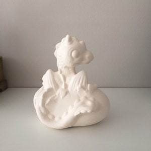 May include: White ceramic dragon figurine sitting on a nest. The dragon has wings and a curled tail.