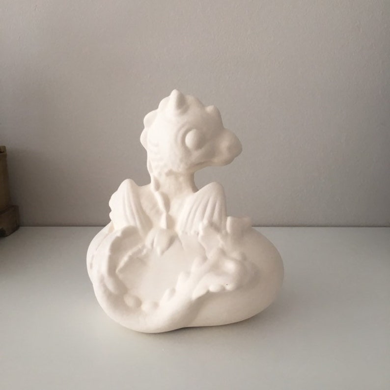 May include: White ceramic dragon figurine sitting on a nest. The dragon has wings and a curled tail.
