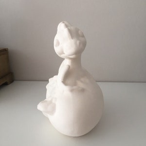 May include: White ceramic figurine of a small dinosaur with a rounded body and a long neck. The dinosaur is sitting with its head tilted up.