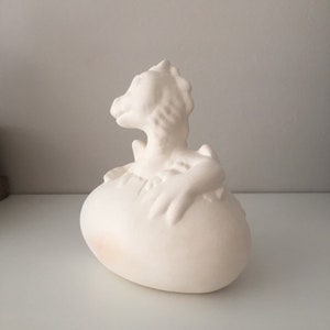 May include: White ceramic dragon sculpture emerging from an egg. The dragon is in a curled position with its head turned to the side. The egg is smooth and oval-shaped.