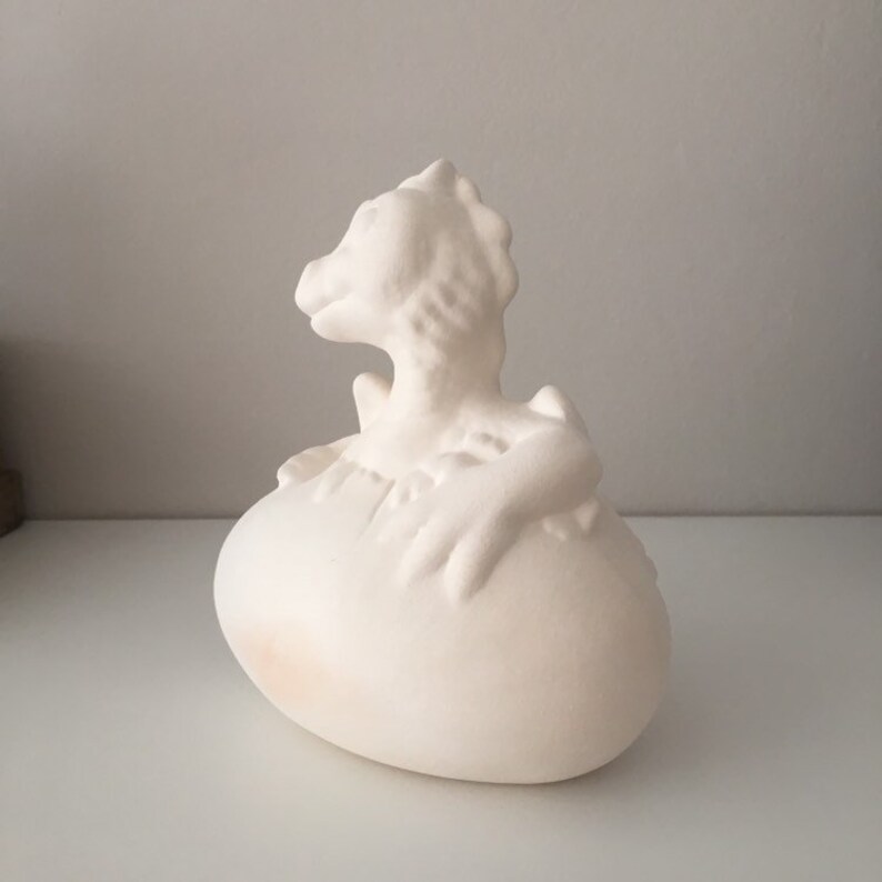 May include: White ceramic dragon sculpture emerging from an egg. The dragon is in a curled position with its head turned to the side. The egg is smooth and oval-shaped.