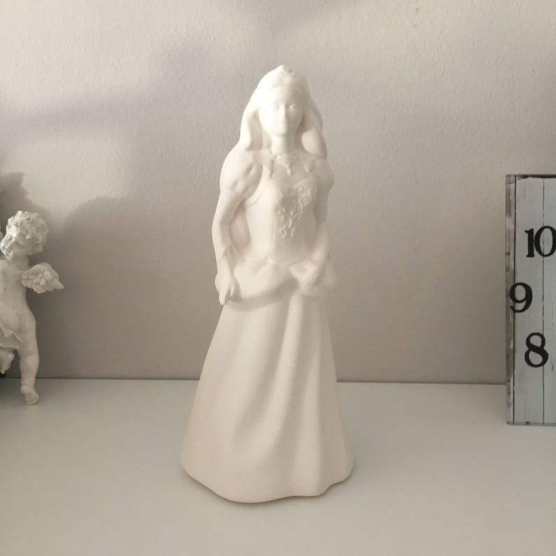May include: White ceramic figurine of a woman wearing a dress and crown. The figurine is standing on a white surface.