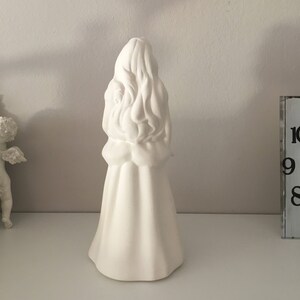 May include: White ceramic figurine of a woman with long hair, wearing a dress. The figurine is standing on a white surface.