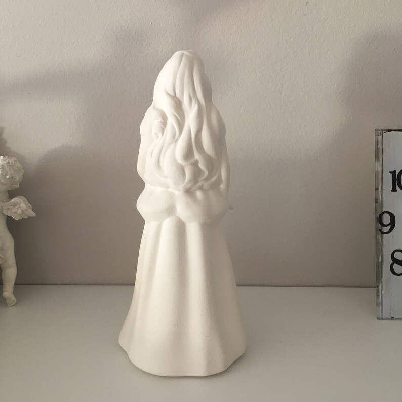 May include: White ceramic figurine of a woman with long hair, wearing a dress. The figurine is standing on a white surface.