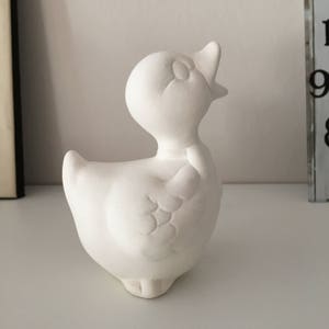 Può includere: Figurina di ceramica bianca di un'anatra con la testa inclinata all'indietro e il becco aperto. L'anatra ha un'ala testurizzata e un piccolo papillon.