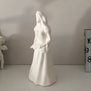 May include: White ceramic figurine of a woman in a long dress with her hair pulled back. The figurine is standing on a white surface.