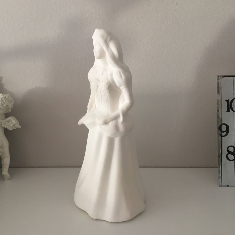 May include: White ceramic figurine of a woman in a long dress with her hair pulled back. The figurine is standing on a white surface.