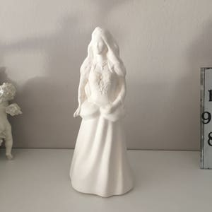 May include: White ceramic figurine of a woman in a long dress with her hair pulled back. The figurine is standing on a white surface.