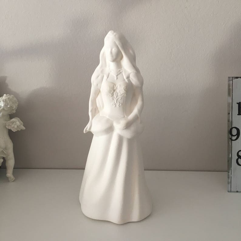 May include: White ceramic figurine of a woman in a long dress with her hair pulled back. The figurine is standing on a white surface.