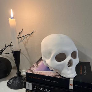 Puede incluir: Una figura de calavera blanca descansa sobre una pila de libros, incluyendo "Dracula" y "The Mysteries of Udolpho". Una vela blanca encendida en un candelabro negro y ramas decorativas completan la escena gótica.