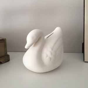 Può includere: Un salvadanaio in ceramica bianca opaca a forma di cigno con una finitura liscia e non smaltata. Il cigno è raffigurato in posizione di riposo con il collo curvato in avanti e una stretta fessura per le monete situata sul dorso dell'uccello.