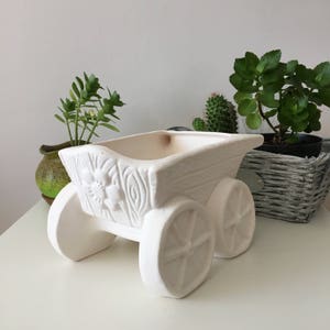 May include: White ceramic planter shaped like a wagon with wheels. The planter has a floral design and is perfect for small plants.