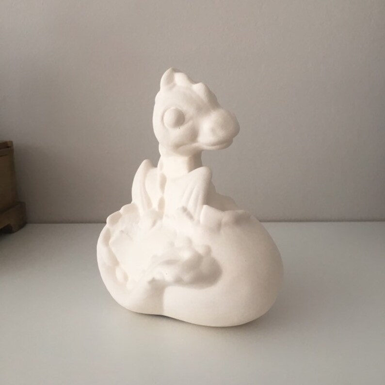May include: White ceramic dragon figurine, hatching from an egg. The dragon has a detailed face and wings, and the egg is textured.