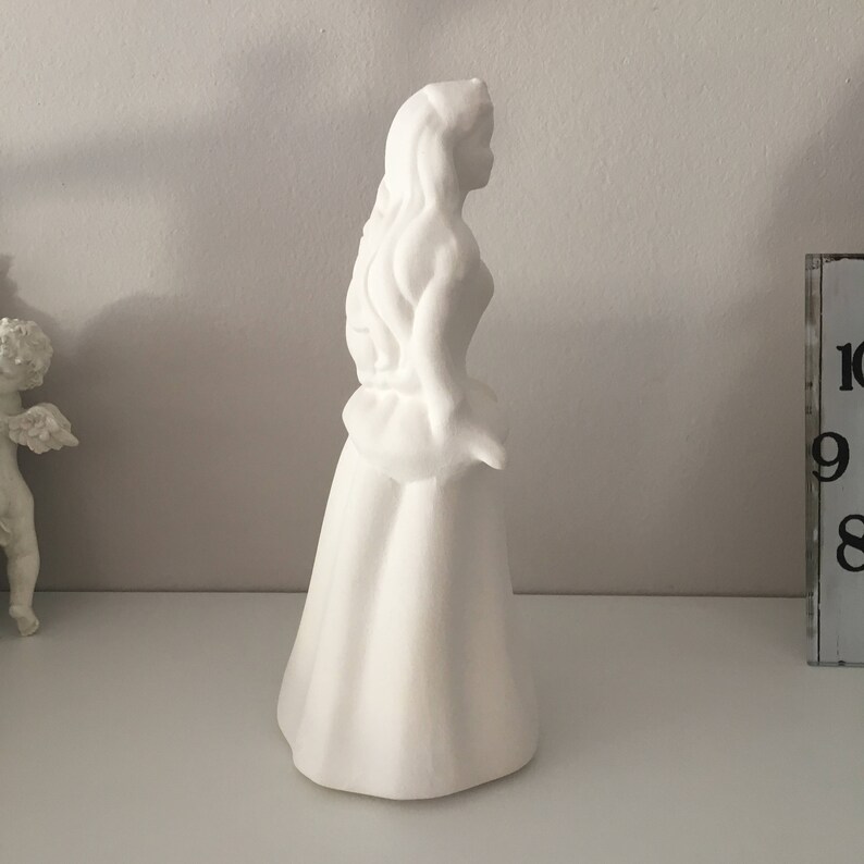 May include: White ceramic figurine of a woman with long hair and a flowing dress. The figurine is standing on a white surface.