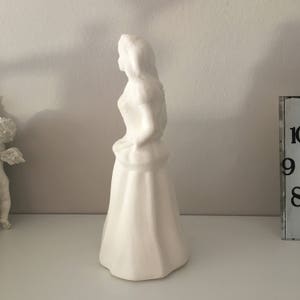 May include: White ceramic figurine of a woman in a long dress. The figurine is standing on a white surface.