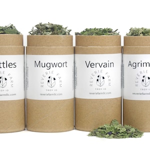 May include: Four brown cardboard tubes with white labels containing dried herbs. The labels read "Nettles", "Mugwort", "Vervain", and "Agrimony". Each label also includes the text "Reverie Farm" and a website address.