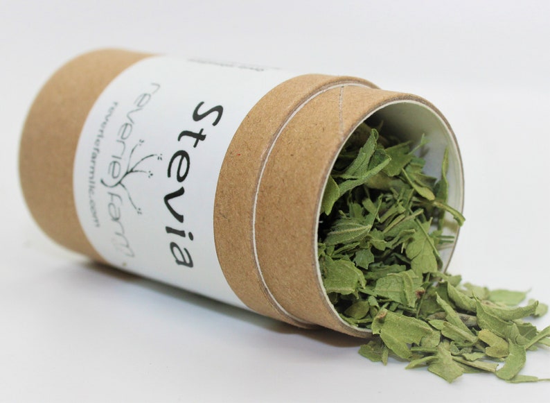 May include: A brown cardboard tube with a white label that says "Stevia" and "Reverie Farm" with a small plant graphic. The tube is open and green dried stevia leaves are spilling out.