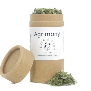 May include: A brown cardboard tube with a white label that says "Agrimony" and "Reverie Farm" with a small illustration of a flower. The tube is open and dried green herbs are spilling out.