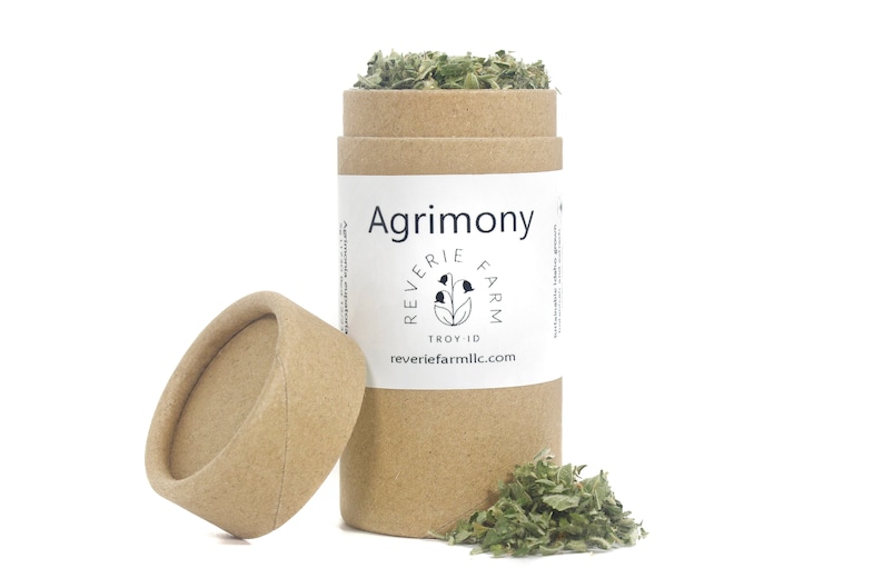 May include: A brown cardboard tube with a white label that says "Agrimony" and "Reverie Farm" with a small illustration of a flower. The tube is open and dried green herbs are spilling out.