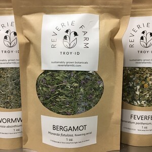 May include: Three brown paper bags with white labels. Each bag contains dried herbs and has a label with the name of the herb, the botanical name, and the weight. The labels also include the name "Reverie Farm" and the location "Troy, ID". The herbs are wormwood, bergamot, and feverfew.