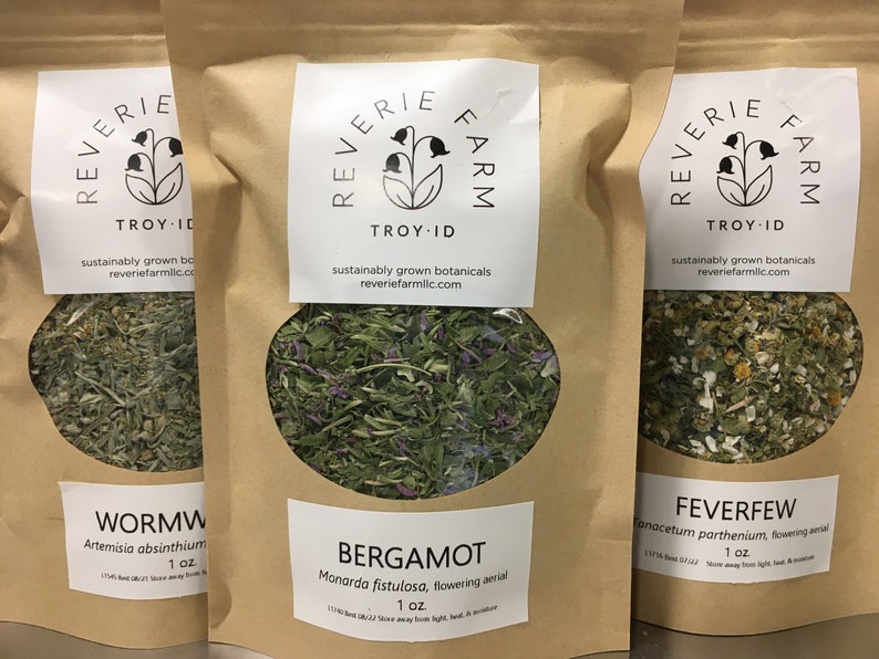 May include: Three brown paper bags with white labels. Each bag contains dried herbs and has a label with the name of the herb, the botanical name, and the weight. The labels also include the name "Reverie Farm" and the location "Troy, ID". The herbs are wormwood, bergamot, and feverfew.