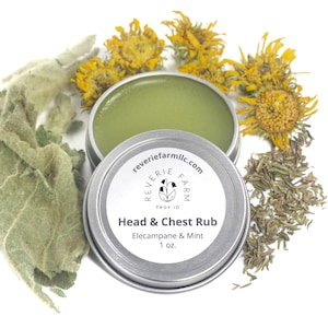 May include: A silver tin with a white label that reads "reveriefarmlic.com", "Reverie Farm", "Troy ID", "Head & Chest Rub", "Elecampane & Mint", and "1 oz.". The tin is filled with a green balm and surrounded by dried herbs.