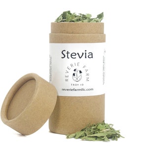 May include: A brown cardboard tube with a white label that says "Stevia" and "Reverie Farm" with a small flower logo. The tube is open and filled with dried green stevia leaves. The lid is off and lying next to the tube.