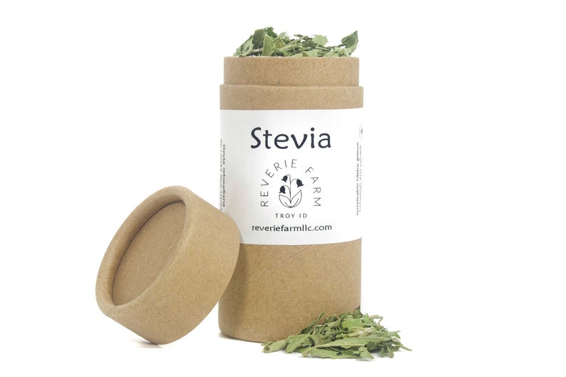 May include: A brown cardboard tube with a white label that says "Stevia" and "Reverie Farm" with a small flower logo. The tube is open and filled with dried green stevia leaves. The lid is off and lying next to the tube.