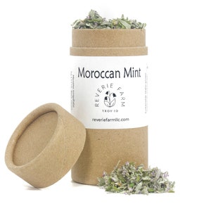 Puede incluir: Un tubo de cart&oacute;n marr&oacute;n con una etiqueta blanca que dice "Moroccan Mint" y "Reverie Farm" con un logotipo de una flor estilizada. El tubo est&aacute; abierto y hay un mont&oacute;n de hierbas secas verdes y moradas delante de &eacute;l.