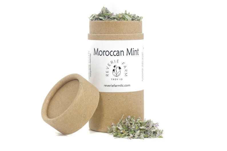 Puede incluir: Un tubo de cart&oacute;n marr&oacute;n con una etiqueta blanca que dice "Moroccan Mint" y "Reverie Farm" con un logotipo de una flor estilizada. El tubo est&aacute; abierto y hay un mont&oacute;n de hierbas secas verdes y moradas delante de &eacute;l.