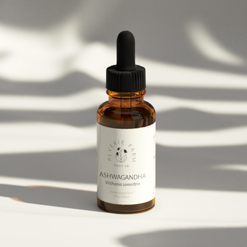 May include: A brown glass bottle with a black dropper top and a white label. The label reads "Reverie Farm Troy ID Ashwagandha Withania somnifera Herbal Supplement 1 fl oz (30ml)"