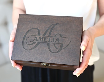 May include: A dark brown wooden box with the name "AMELIA" engraved on the lid. The box is rectangular with a decorative clasp. The lid features a large, stylized letter "A" above the name.