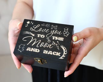 May include: A black wooden box with the phrase "Love you to the Moon and Back" engraved on the lid. The box has a metal clasp and is held by a person. The text is in a cursive font with star and comet illustrations.