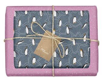 Può includere: Una scatola regalo rettangolare avvolta in carta rosa con macchie bianche, con un pannello centrale di carta blu scuro con un motivo a pinguini e linee geometriche bianche. Il pacchetto è legato con spago marrone e un cartellino marrone.
