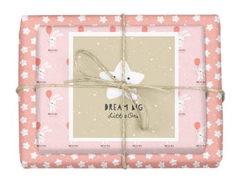 Könnte beinhalten: Ein rechteckiges Geschenkpaket, eingewickelt in rosa Papier mit einem wiederkehrenden Muster aus weißen Sternen und Hasen, die rote Luftballons halten. Eine beige quadratische Karte mit einem weißen Stern und dem Text "DREAM BIG Little One" ist mit Bindfaden befestigt.