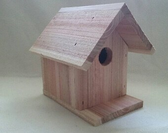 Puede incluir: Una pequeña casa para pájaros de madera clara con un tejado a dos aguas y un agujero de entrada circular. La casa para pájaros tiene una base rectangular. El diseño es sencillo y rústico, adecuado para uso en exteriores.