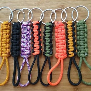 May include: A collection of eight handmade keychains with woven paracord designs in various colors, including yellow, black, purple, orange, green, and black. Each keychain has a silver metal ring and a loop at the bottom.