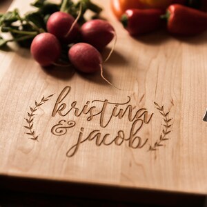 Peut inclure: Une planche &agrave; d&eacute;couper en bois personnalis&eacute;e avec les noms "kristina & jacob" grav&eacute;s en &eacute;criture cursive. La planche est entour&eacute;e d'un motif de couronne d&eacute;coratif.