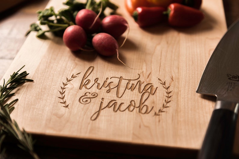 Peut inclure: Une planche &agrave; d&eacute;couper en bois personnalis&eacute;e avec les noms "kristina & jacob" grav&eacute;s en &eacute;criture cursive. La planche est entour&eacute;e d'un motif de couronne d&eacute;coratif.