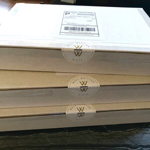 May include: Three white cardboard boxes stacked on top of each other. Each box has a gold and white sticker with the words "Well Written Gifts" and a stylized "W" logo.