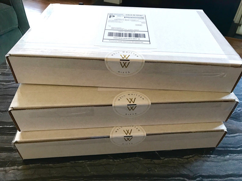 May include: Three white cardboard boxes stacked on top of each other. Each box has a gold and white sticker with the words "Well Written Gifts" and a stylized "W" logo.