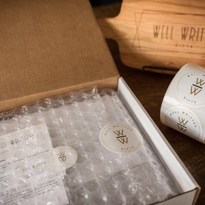 May include: A brown cardboard box with a white interior is open and filled with bubble wrap. The box is labeled "Well Written Gifts". There are also several round white stickers with the same logo on them.