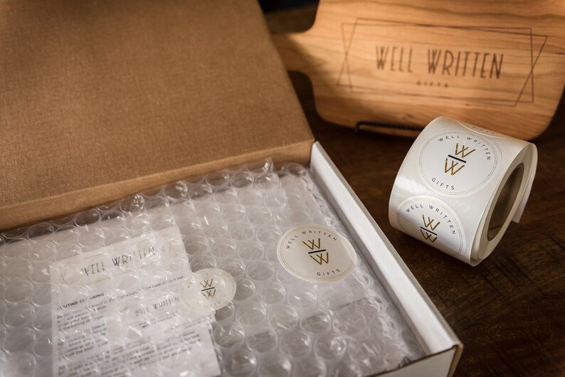 May include: A brown cardboard box with a white interior is open and filled with bubble wrap. The box is labeled "Well Written Gifts". There are also several round white stickers with the same logo on them.
